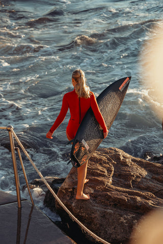 Person in a red wetsuit holding a surfboard on a rocky outcrop with ocean waves in the background